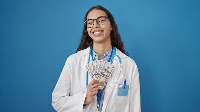 Young beautiful hispanic woman doctor smiling holding money over isolated blue background