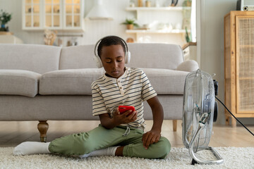 Interested african american child boy sit with smartphone in headphones on floor pleasure cooling with electric fan. Satisfied kid schoolboy use cellphone listen music chilling at home. Overheating. 