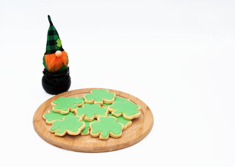 St. Patrick's Day green iced shamrock cookies on a round wooden platter are being watched by a toy leprechaun sitting in a black cauldron against a white background.