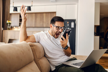 An angry man sits at home on sofa and yelling at the phone while holding a laptop in his lap.