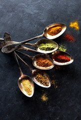 Spices, herbs and seasonings in spoons on a black textured background, flat lay
