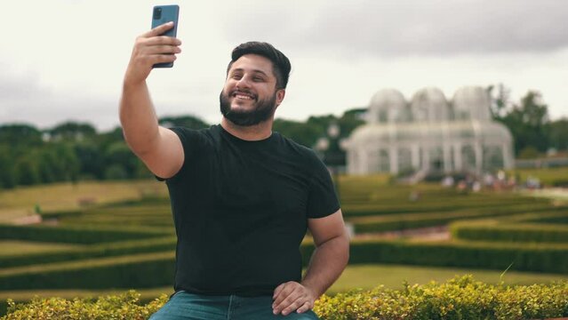 Young handsome man picking up his cell phone and taking a selfie at a park outdoors while seated on a bench located at Botanical Garden, Curitiba, Brazil	