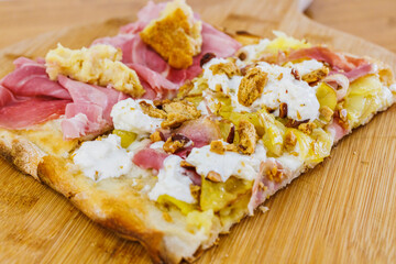 Due fette di Pinsa Romana condite con con pancetta, burrata, patate e tarallo sbriciolato e con prosciutto cotto, crocchè di patate e mozzarella poggiate su un tagliere di legno