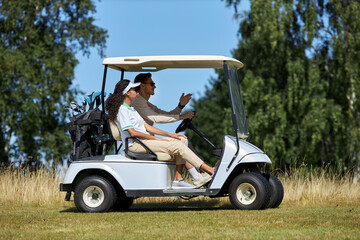 Side view portrait of sporty young couple driving golf cart across field in sports club lit by...