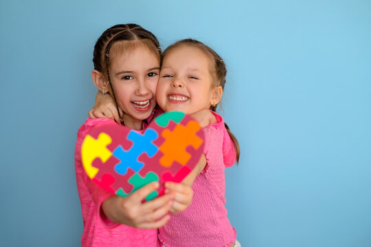 Funny laughable girls show a heart- shaped postcard with pieces of multicolored puzzles . Heart in Viva Magenta color for children with autism