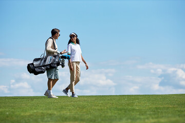 Minimal side view portrait of elegant sporty couple carrying golf bag walking on green field against blue sky, copy space - Powered by Adobe