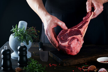 Chef cutting steak beef. Mans hands hold raw steak Tomahawk on rustic wooden cutting board on black...