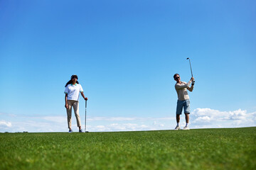 Full length portrait of active sporty couple playing golf on field against blue sky, copy space