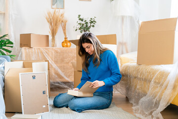 Young female sitting on floor near carton boxes and reading notes in clipboard