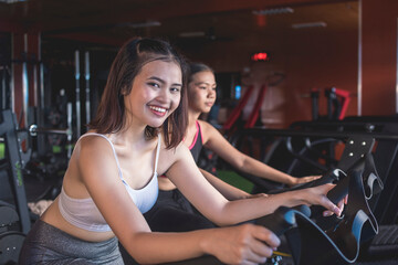 Fototapeta na wymiar Two young asian women doing warmup cardio on a stationary bike. 2 friends working out together at the gym.
