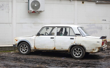 A white rusty old car is parked near a white plastic wall, Antova-Ovseenko Street, St. Petersburg, Russia, March 2023