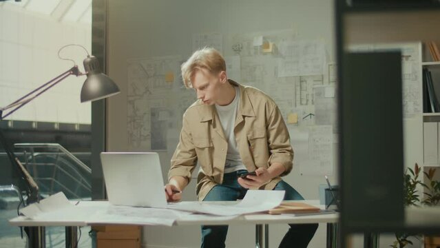 Handsome Creative Designer or Architect Working on Project in Office Using a Laptop Computer and Phone with Drawings,Handsome Focused Man Doing Project Sitting at Workplace Indoors Sketching a Plan
