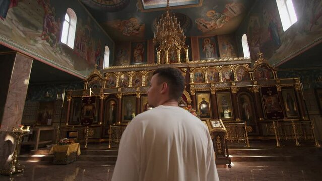 Young man stands in front of iconostasis and examines the rich decoration of orthodox church with frescoes. Back view