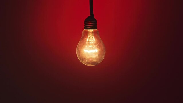 Incandescent lamp glows and flickers on dark red background. Edison light bulb slowly turned on and off, pulsing light close-up. Warm flashing filament. Retro classic bulb. Zoom out