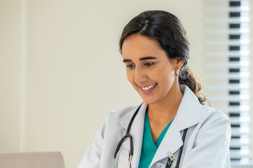 Woman female doctor portrait in white coat gown with stethoscope sit and smile to front. Medical student attractive friendly wear white coat smiling and work on table in patient room