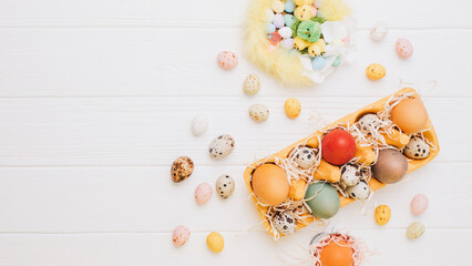 Painted multi-colored eggs in a yellow cardboard tray on a white wooden background. Easter background, web banner. Top view