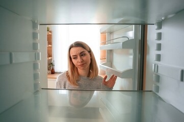 Fototapeta na wymiar Disappointed about an empty fridge with no food in it. Beautiful young woman is on the kitchen