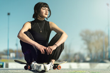 Pretty young girl wearing glasses sitting on her skateboard in the floor towards the sunlight