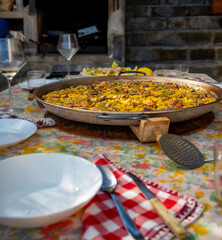 Paellera with a freshly cooked authentic Valencian paella, to serve on a table with the dishes set, salad, glasses and the floral tablecloth..