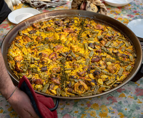 Chef holding a paella pan with a freshly cooked authentic Valencian paella, to serve on a table with the plates set and the tablecloth.
