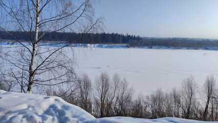 Winter landscape with trees on a cliff and a view from a height of a frozen river or a field with snow on a cold sunny day with a blue sky