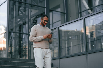 Walking, holding smartphone. Handsome black man is outdoors near the business building