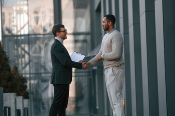 Doing handshake, greetings gesture. Businessman in formal clothes is with black man outdoors in the city © standret