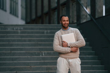 Positive handsome black man is outdoors near the business building holding laptop