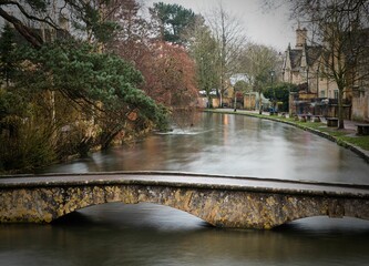 Fototapeta premium Long exposure of the river through Bourton on the Water with trees in The Cotswolds in winter