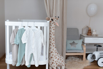 Baby bodysuit on hanger on bed, beige background. Cute children's clothes, baby chair, wooden...