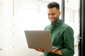 Smiling freelancer guy wearing casual shirt using laptop in contemporary office space, concentrated man typing, messaging, indian programmer develops software, looks at computer screen