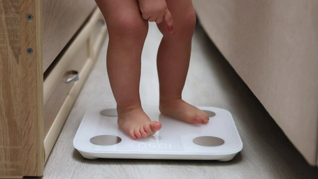 baby legs barefoot feet on body composition scale measuring weight home interior laminate wooden floor background bed and toddler crib.cute pretty fat infant legs on white scale digital numbers