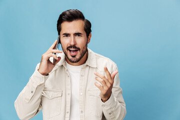 Portrait of a brunette man talking on the phone mobile and internet online, anger and frustration problems, on a blue background in a white T-shirt, copy space