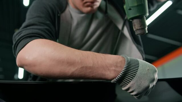 A worker covers a car spoiler with a film in the workshop using a hair dryer and a spatula close-up Car detailing and care