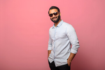 Obraz premium Handsome bearded Indian man wearing sunglasses in casual jeans shirt looking at camera and smiling isolated on pink. Charismatic middle-eastern guy stands with hands in pockets