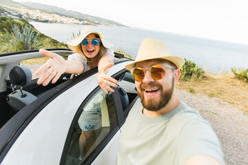 Naklejka premium Romantic couple making selfie on smartphone camera in rental cabrio car on ocean or sea beach enjoying summer vacation together and taking picture on cellular resting near sea on weekends