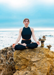 young female is sitting in yoga lotus pose on the stone near the ocean