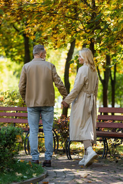Carefree woman in trench coat holding hand of husband while walking in park.