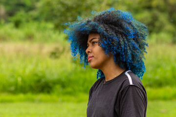 Foto de uma jovem negra de cabelos afro, tingidos de azul, com expressão serena no rosto, de perfil com a vegetação borrada do parque ao fundo