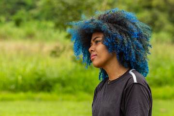 Foto de uma jovem negra de cabelos afro, tingidos de azul, com expressão serena no rosto, de perfil com a vegetação borrada do parque ao fundo - obrazy, fototapety, plakaty