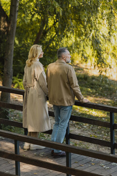 Side view of middle aged couple in spring outfit standing on bridge in park.