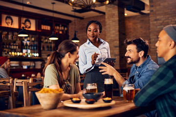 Happy African American waitress takes order from group of guests in pub.
