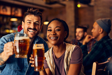 Happy multiracial couple toasting with beer in pub and looking at camera.