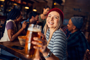 Happy woman toasts with beer while being with friends in bar and looking at camera.