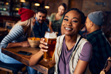 Happy black woman drinking beer with friends in pub and looking at camera.