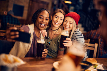 Happy female friends drinking beer and taking selfie in bar.