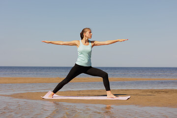 Fototapeta premium Virabhadrasana, woman in sportswear do sports fitness and yoga on seashore. athletic millennial female trains before marathon, healthy lifestyle, love for her body, mental and physical condition, 