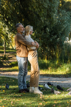 full length of happy middle aged man hugging blonde wife while standing near pigeons in park.