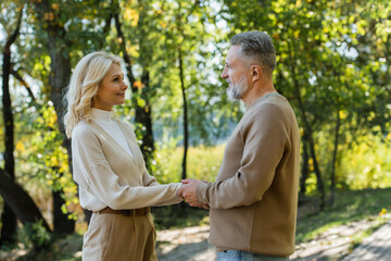 Obraz premium side view of happy mature couple holding hands and looking at each other in green park.