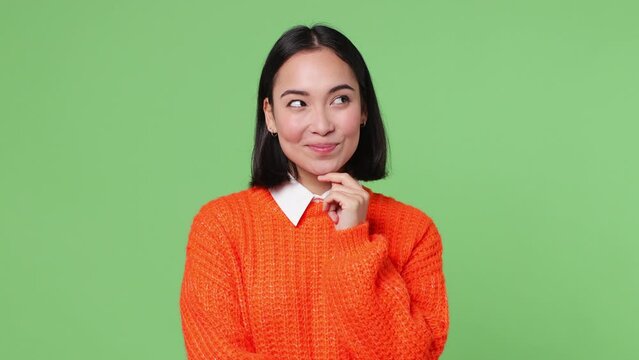 Beautiful young woman of Asian ethnicity she wear orange sweater look around think dream put hand prop up on chin lost in thought and conjectures isolated on plain pastel light green color background